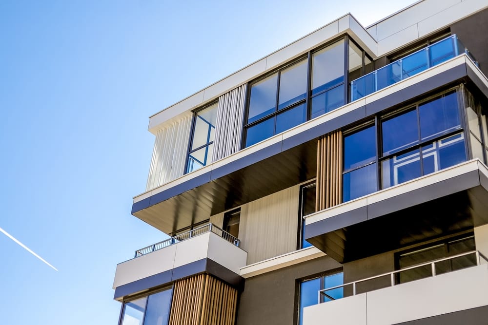 Modern apartment building with large glass windows and wooden accents under a clear blue sky - RM Builders Associates Inc Modern apartment building with large glass windows and wooden accents under a clear blue sky - RM Builders Associates Inc